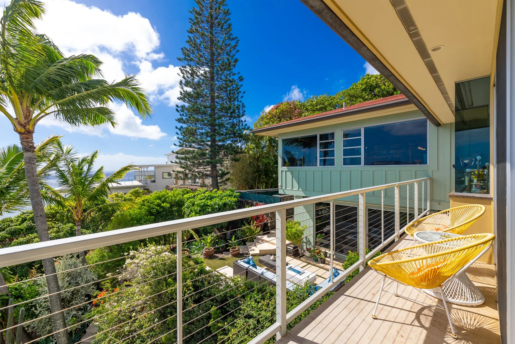 A sunny balcony overlooks a garden, with outdoor seating beside the railing.