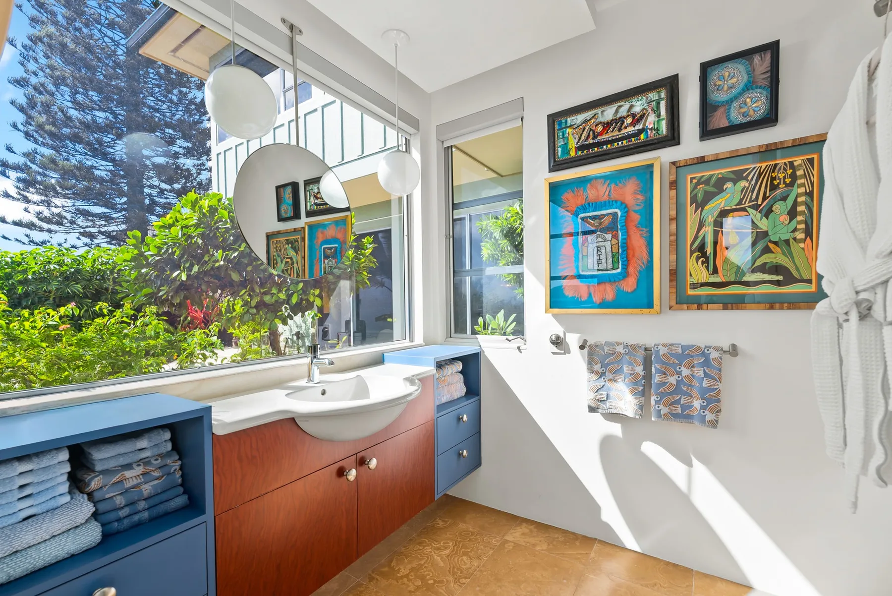 A bright bathroom with a sink, large windows, and colorful framed artwork.