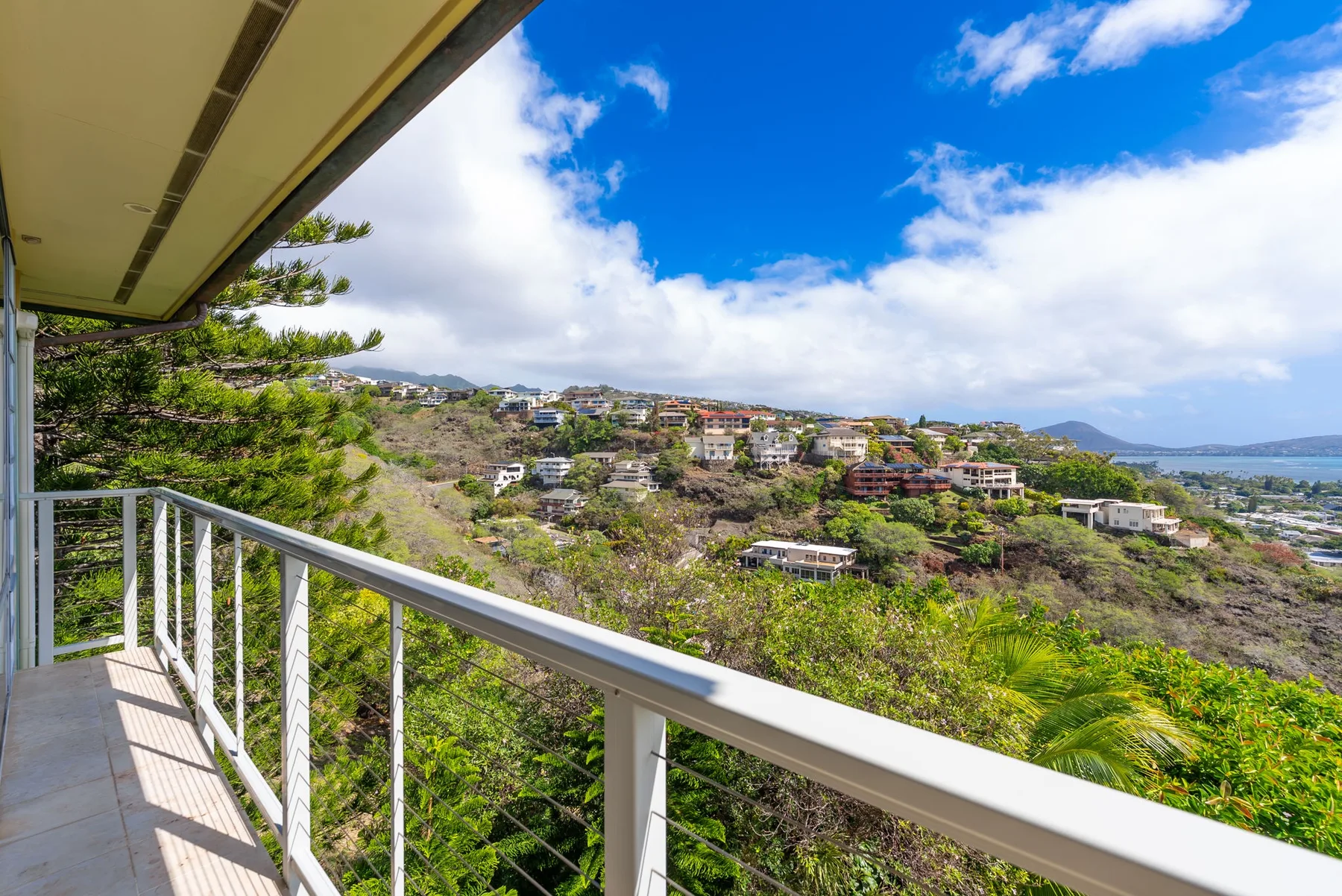 A balcony overlooks a green hillside dotted with homes beneath a partly cloudy sky.