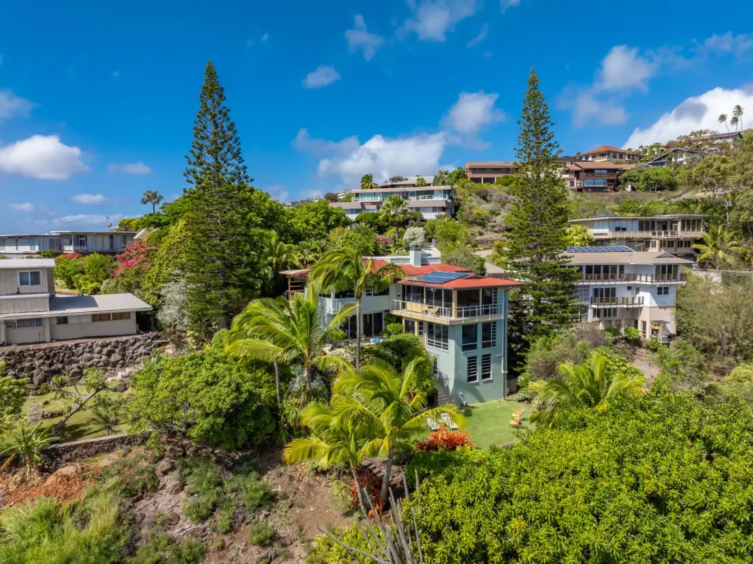 A hillside neighborhood of homes sits among trees beneath a blue sky.