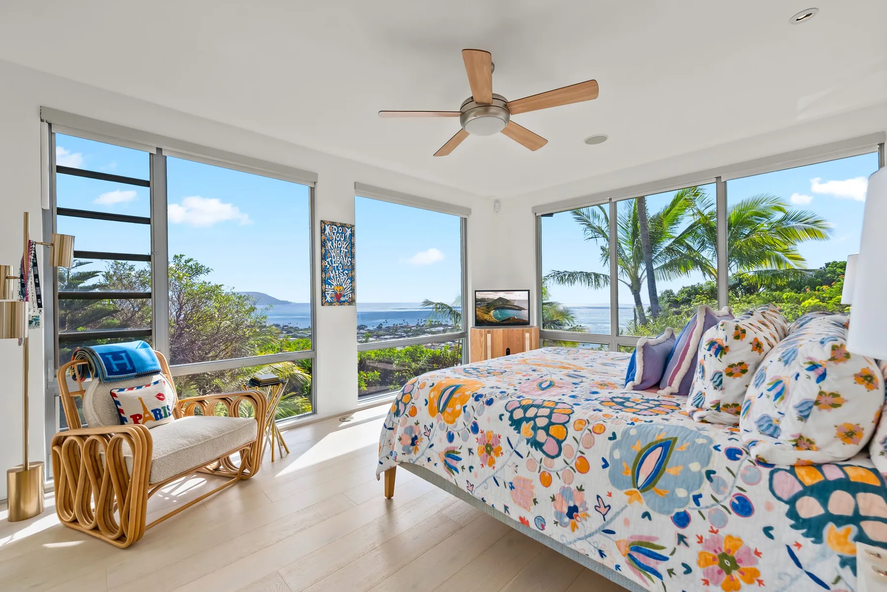 A bright bedroom with a patterned bed and large windows overlooking palm trees.