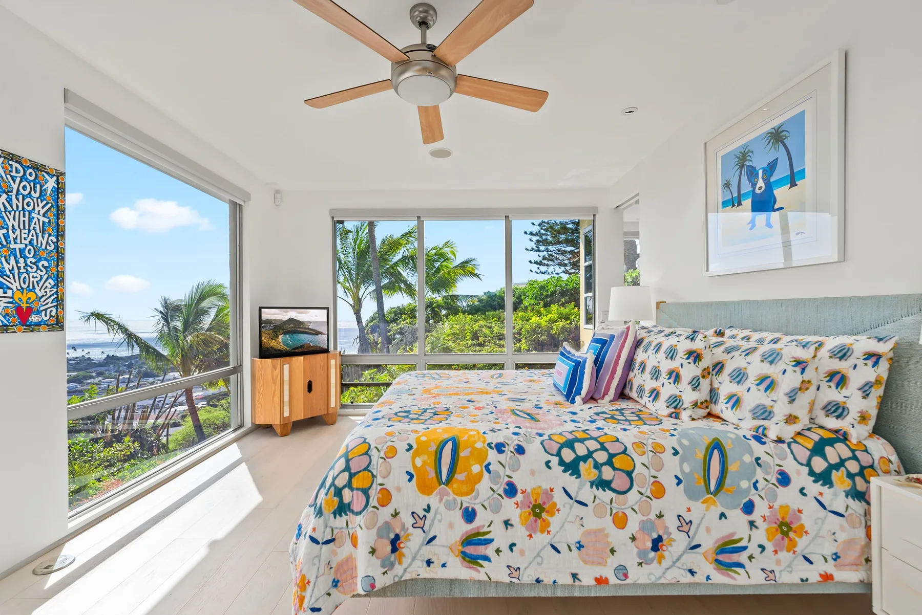 A bright bedroom with a colorful floral bedspread and large windows overlooking palm trees.