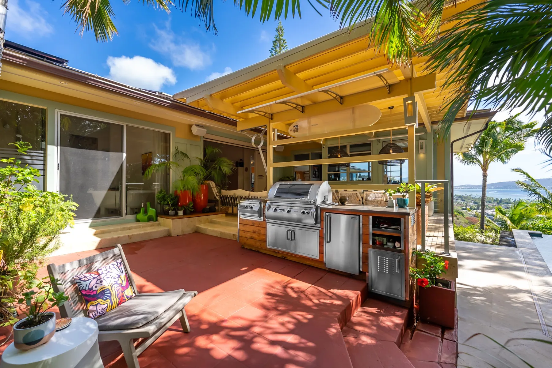An outdoor kitchen and patio beneath a yellow pergola, with plants and seating.