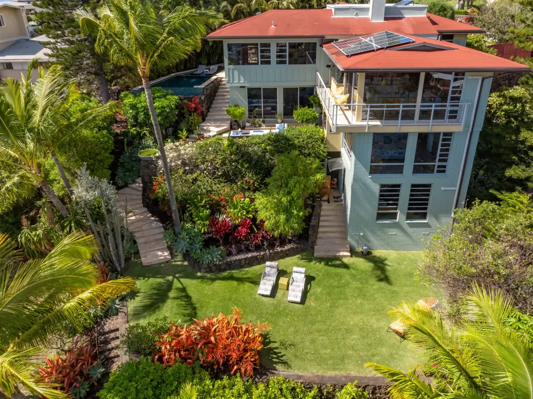 An aerial view shows a modern house surrounded by tropical gardens and lawn.