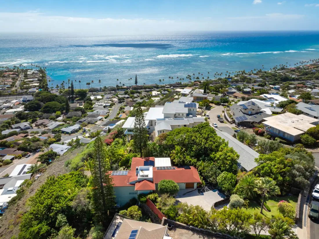 An aerial view shows homes among green trees beside a blue ocean.