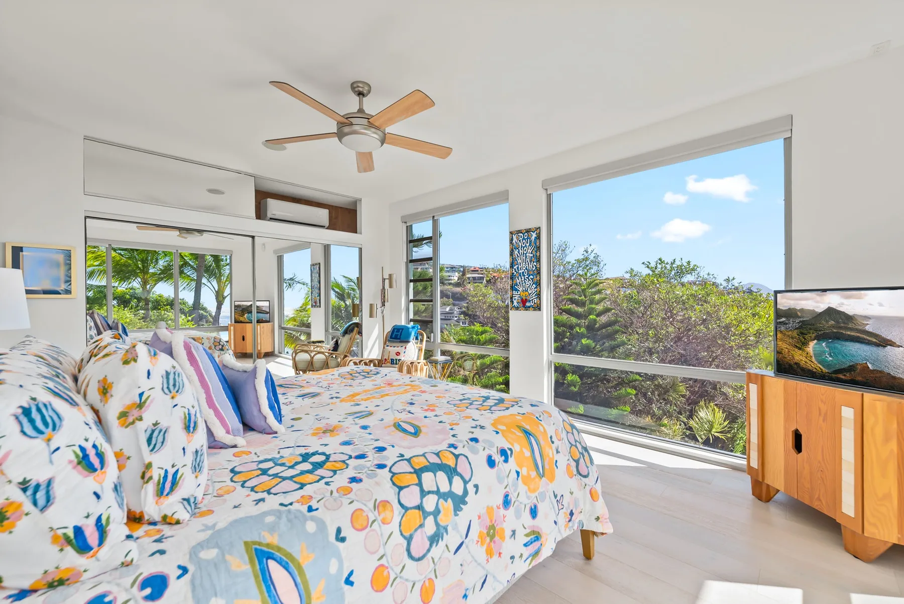 A bright bedroom with a colorful bed beside large windows overlooking greenery.