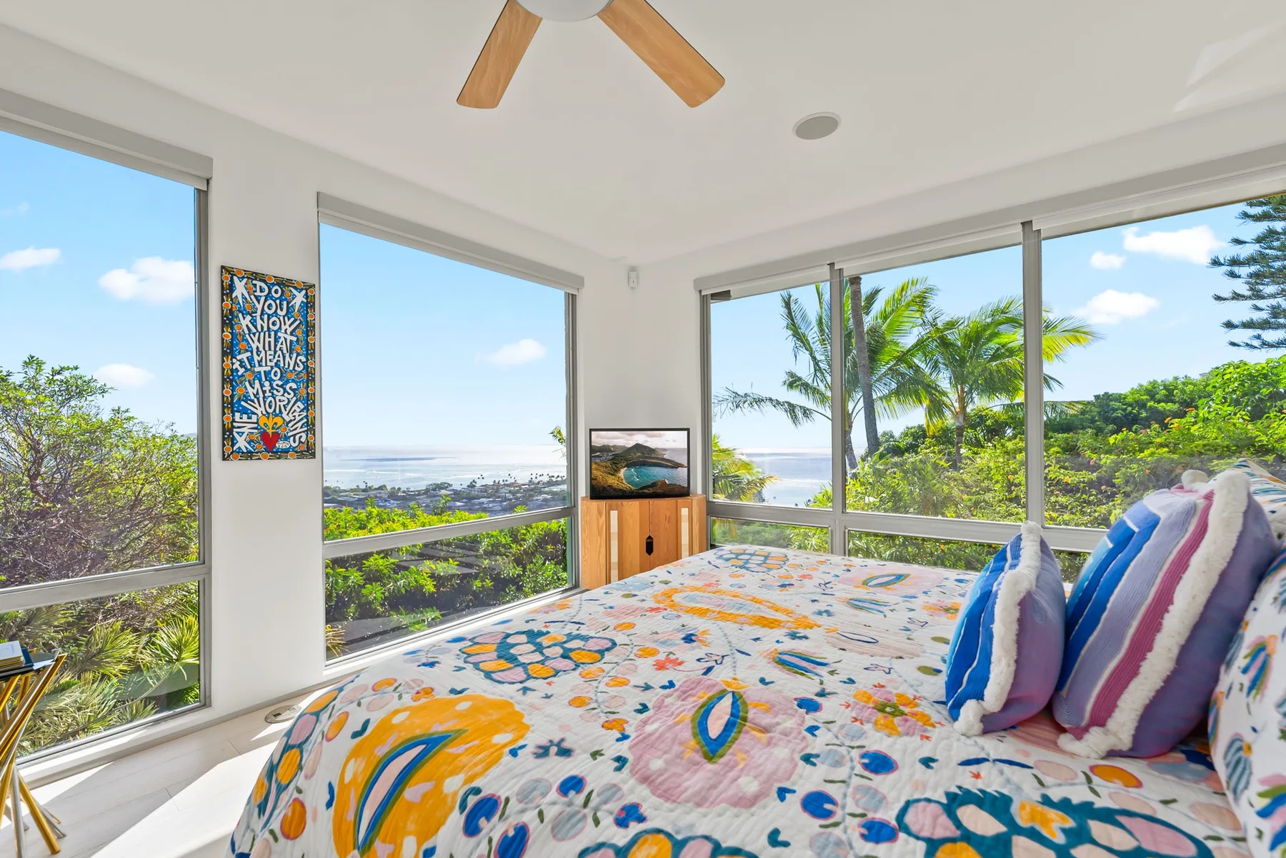 A bright bedroom has a colorful bed beside large windows overlooking palm trees and greenery.