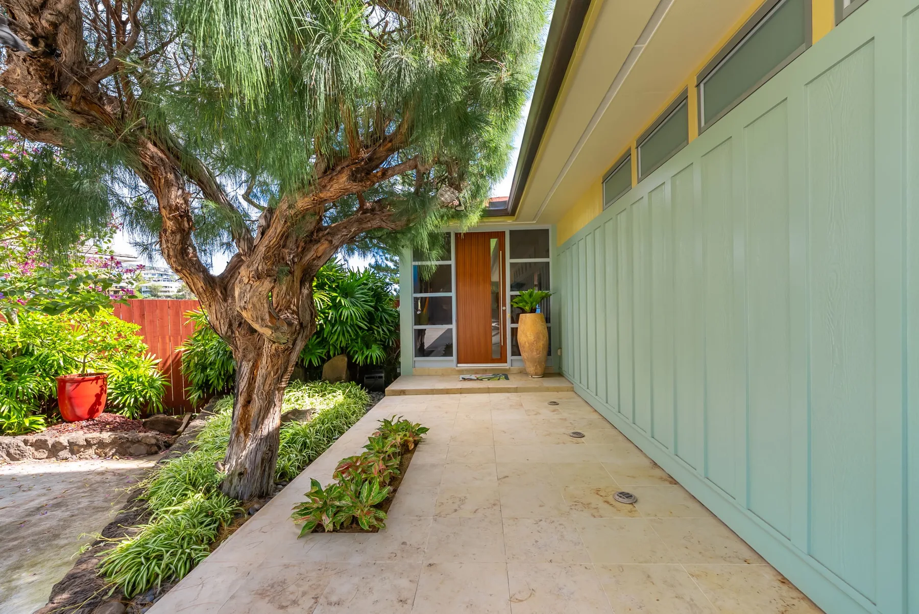 A covered entry walkway beside a leafy garden leads to a glass door.
