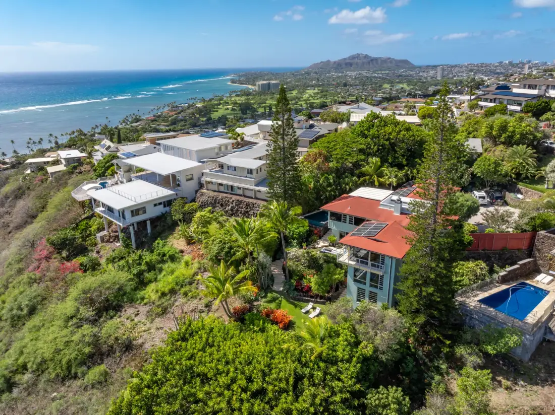 Aerial view of coastal homes among greenery, with the ocean and distant hills.