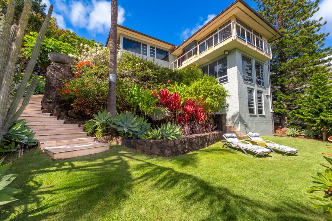 A landscaped garden surrounds a multi-story house, with a lawn and outdoor seating.
