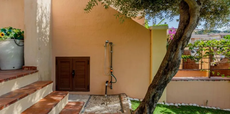 Una ducha exterior está junto a escalones de baldosas y un árbol frondoso.