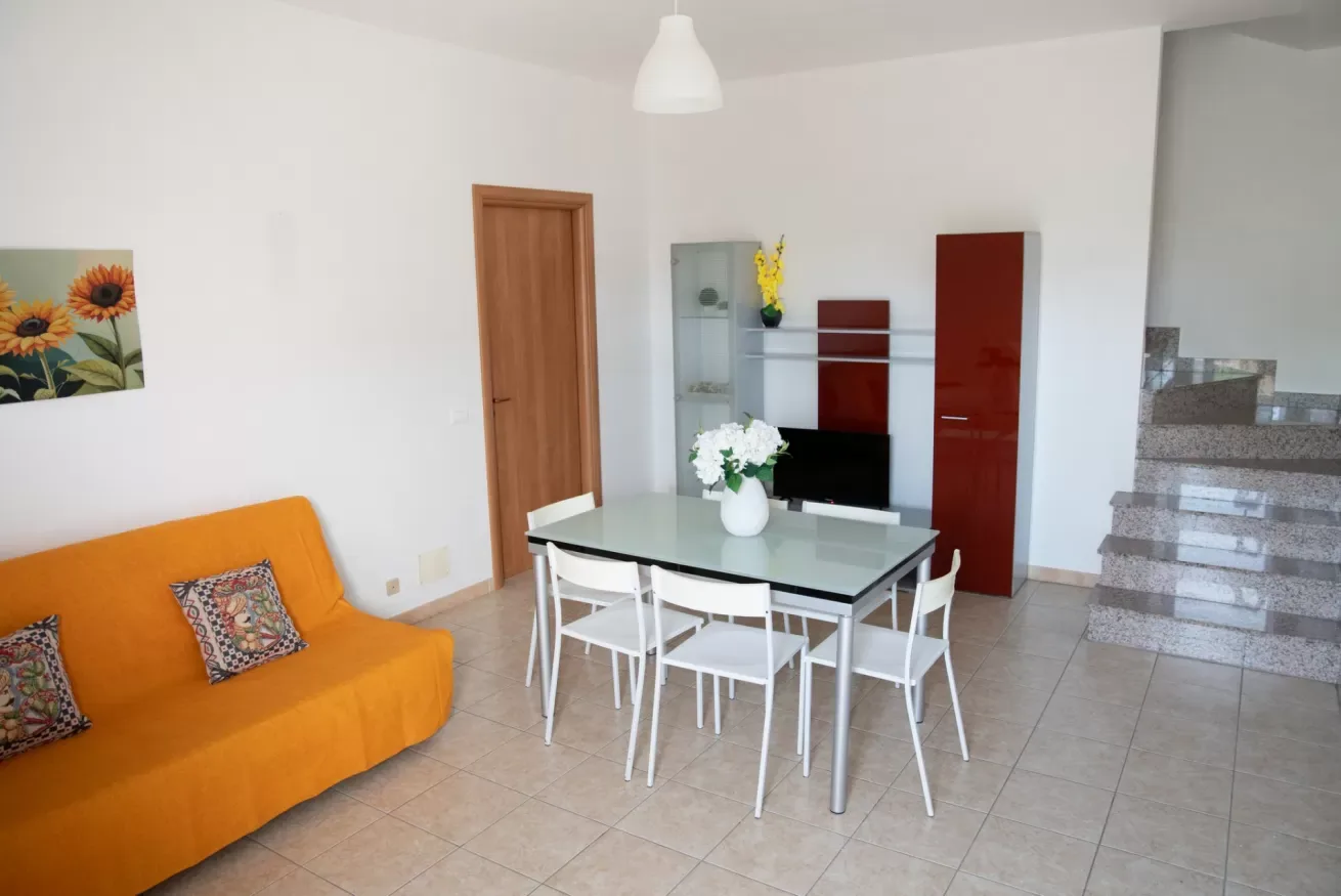 Comedor con una mesa de vidrio y sillas blancas junto a un sofá naranja.