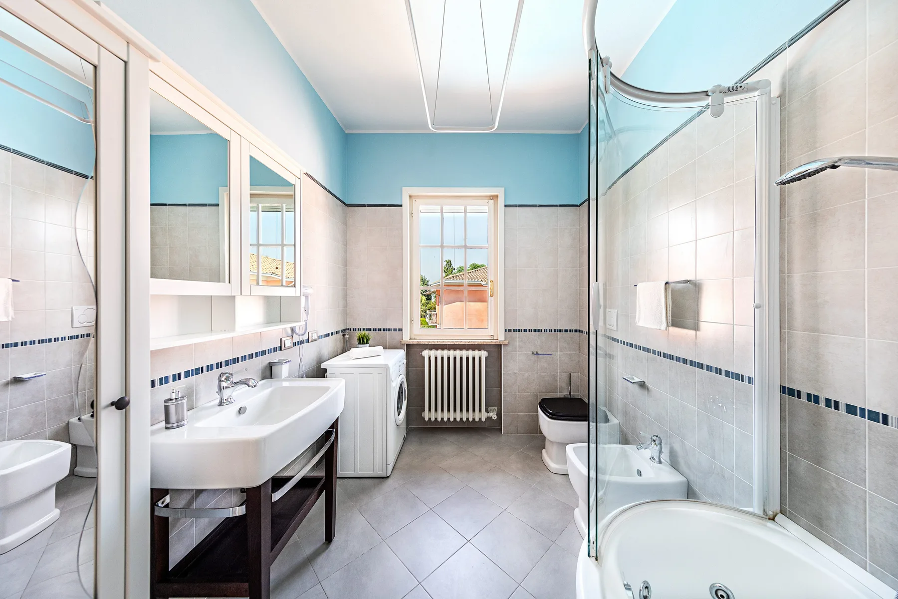 A bright tiled bathroom has a long vanity, a bathtub, and a glass shower screen.