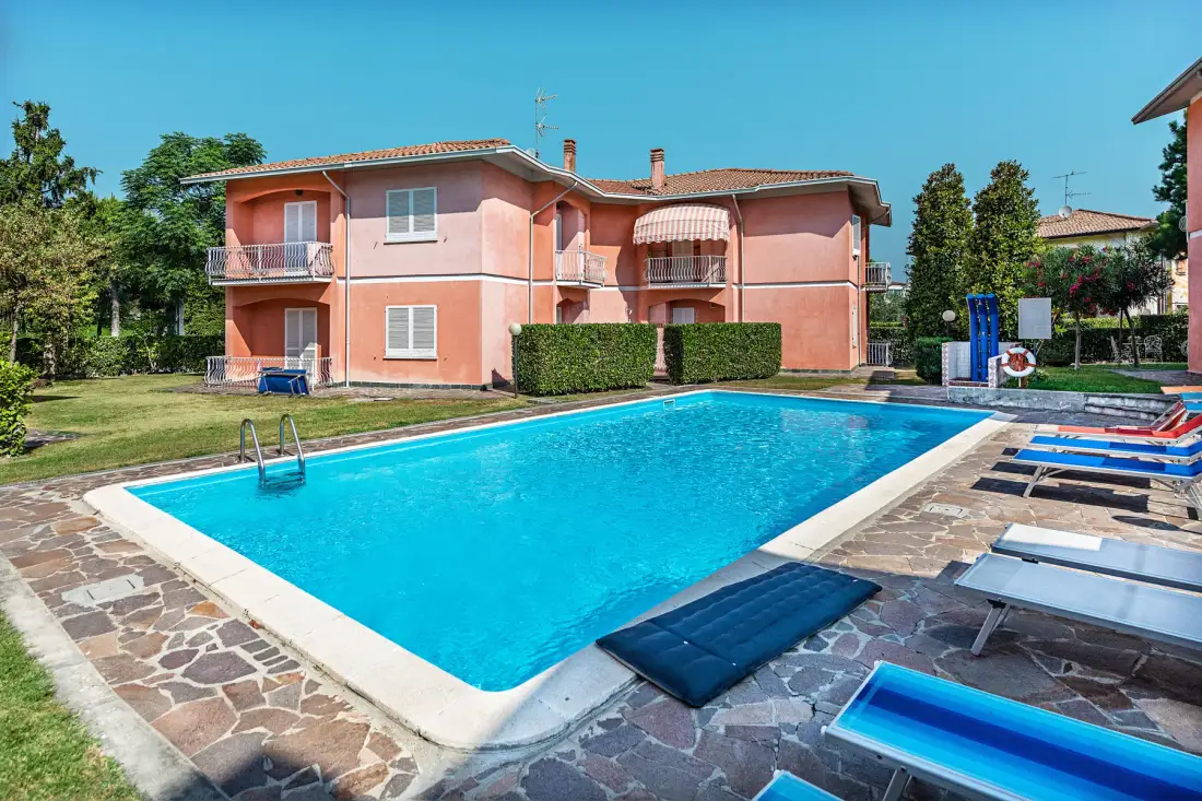 An outdoor swimming pool sits beside a pink residential building and paved deck.
