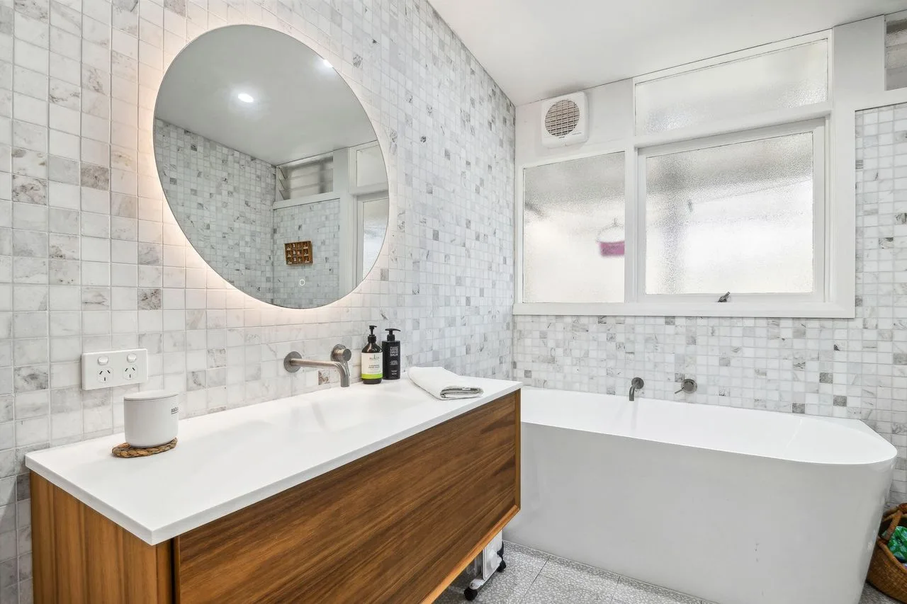 A tiled bathroom with a vanity, round mirror, and bathtub.