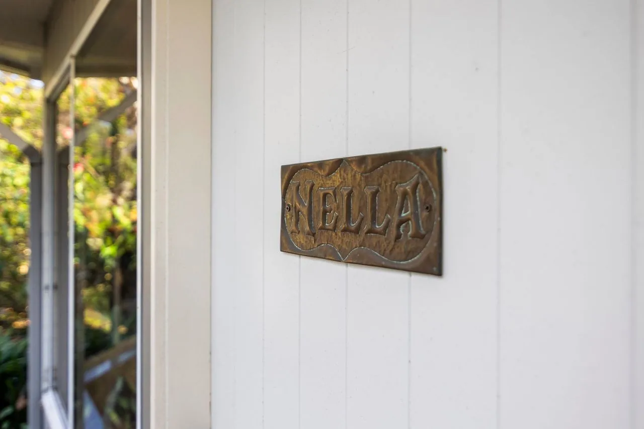 A carved wooden sign reading “Nella” is mounted on a white exterior wall beside a window.