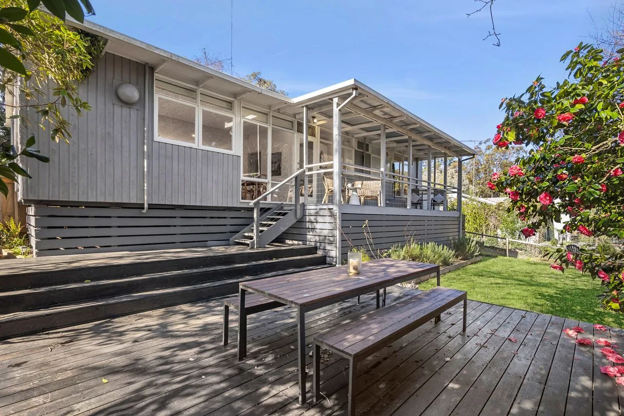 A raised house opens onto a wooden deck with a picnic table and garden.