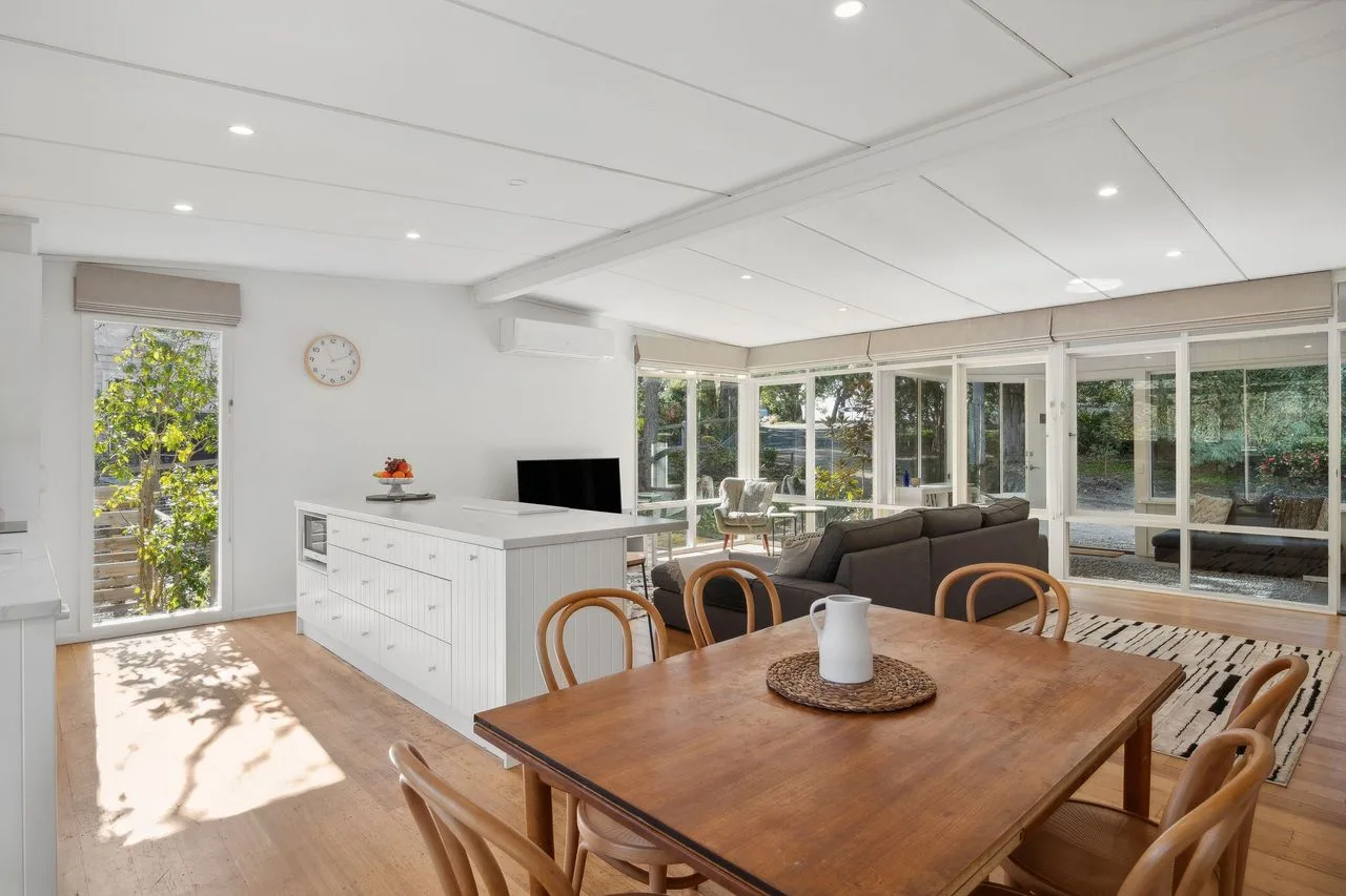 Bright dining and kitchen area with a wooden table beside broad windows.
