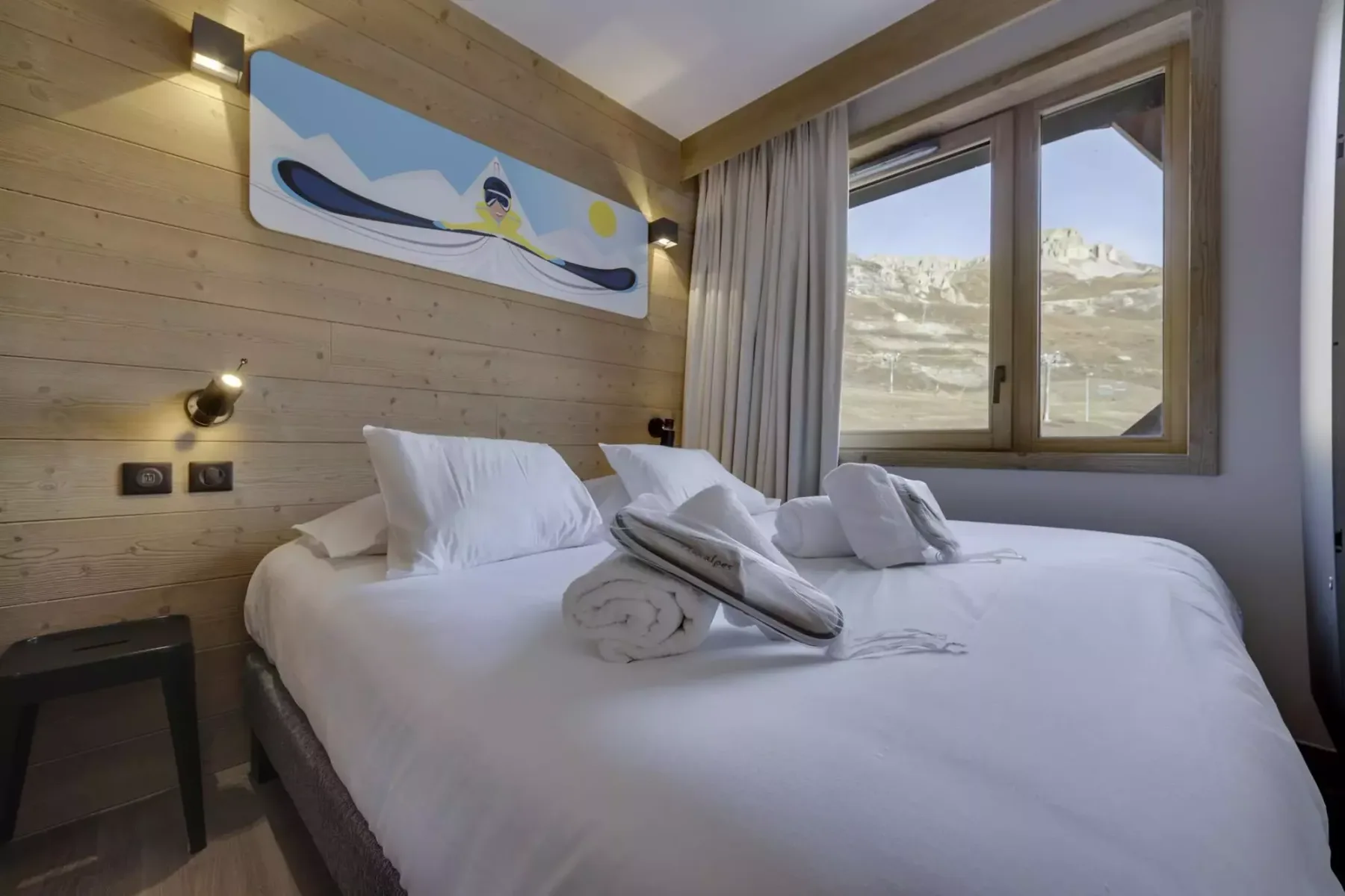 A bedroom with white bedding, folded towels, and a window overlooking a rocky landscape.
