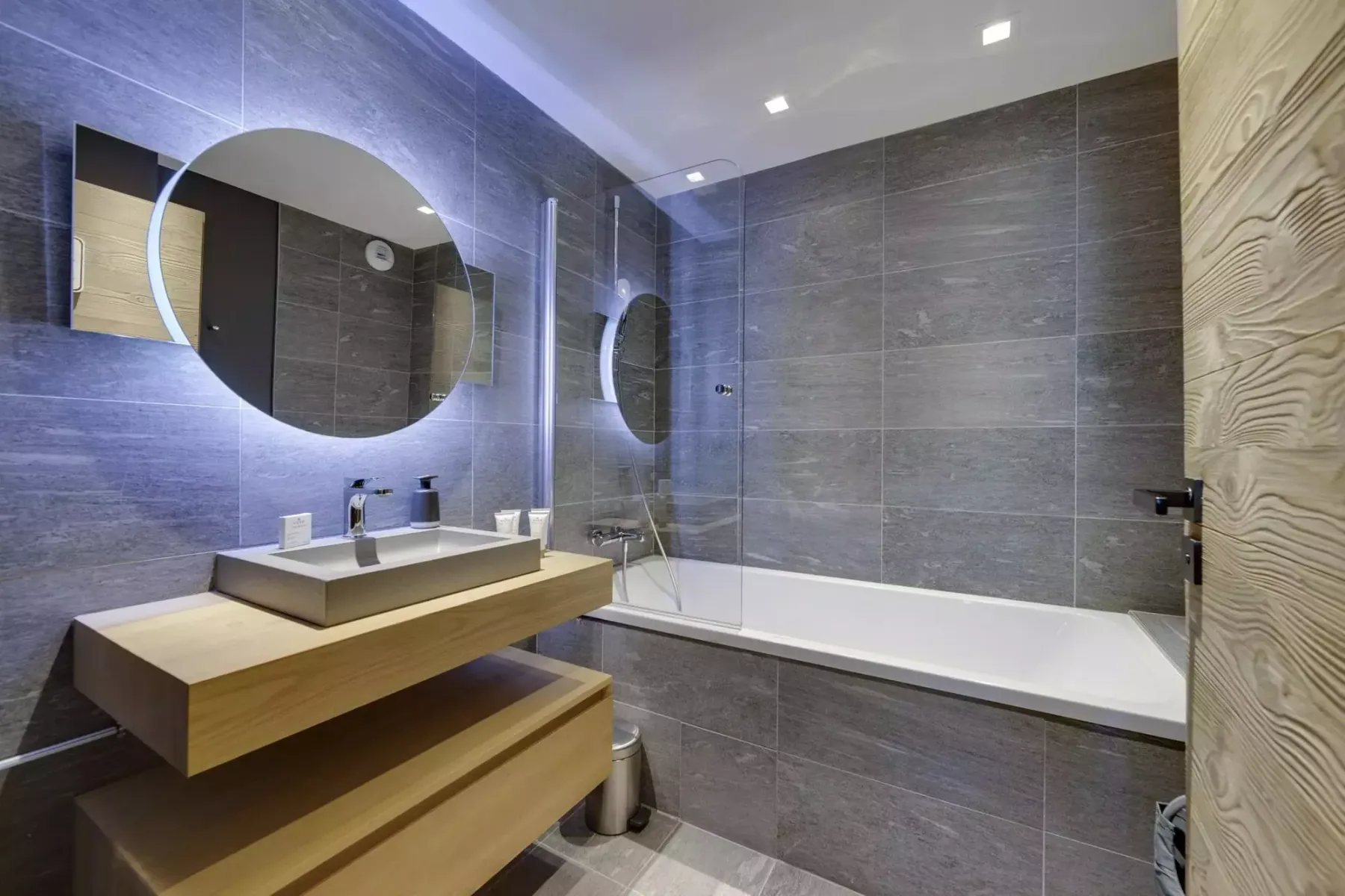 A modern bathroom with a bathtub, wall-mounted shower, and wooden vanity.