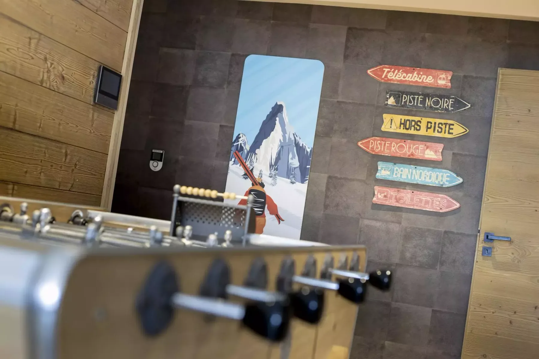 A foosball table stands before a wall with a mountain illustration and colorful signs.