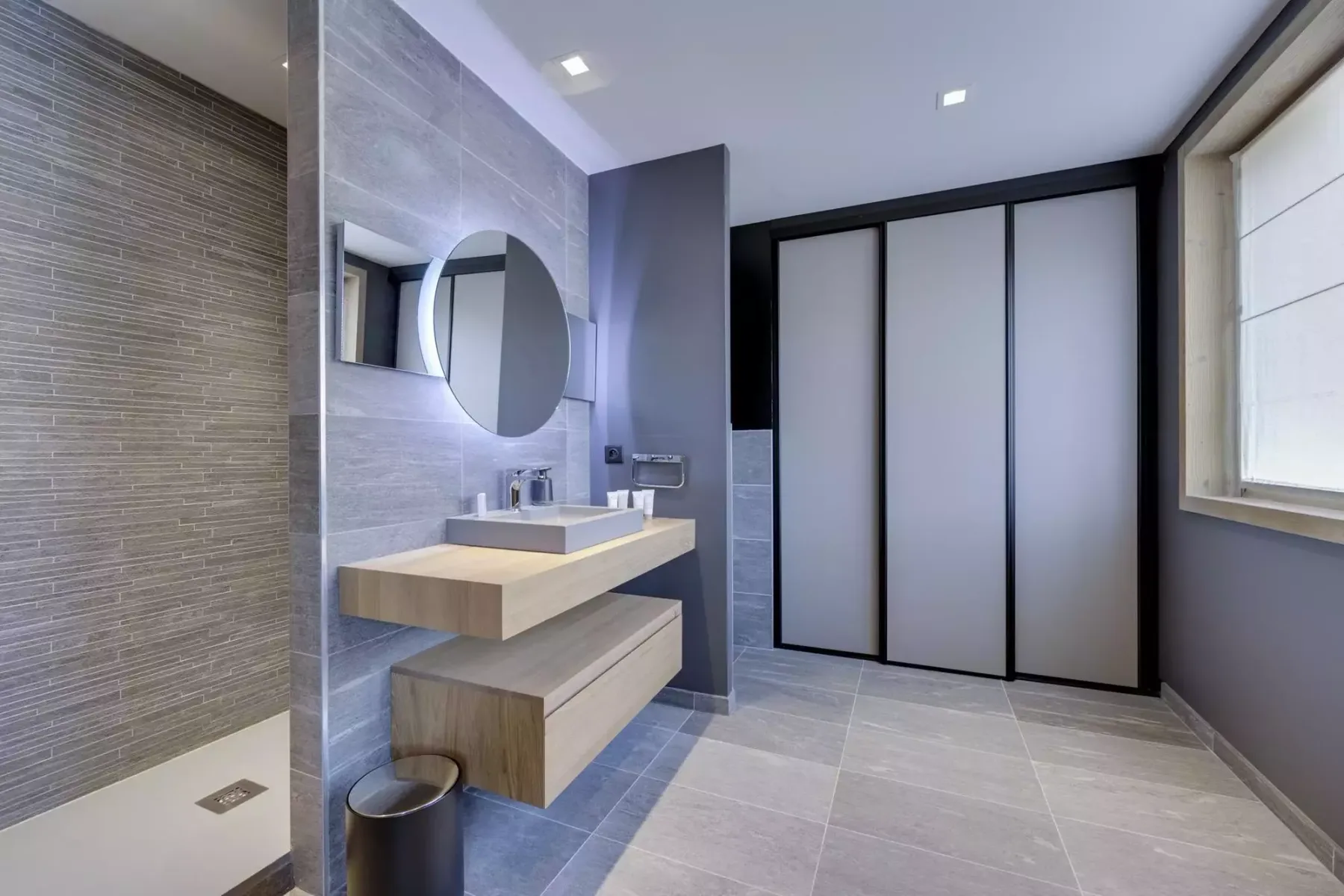 A modern bathroom with a floating vanity, round mirror, and walk-in shower.