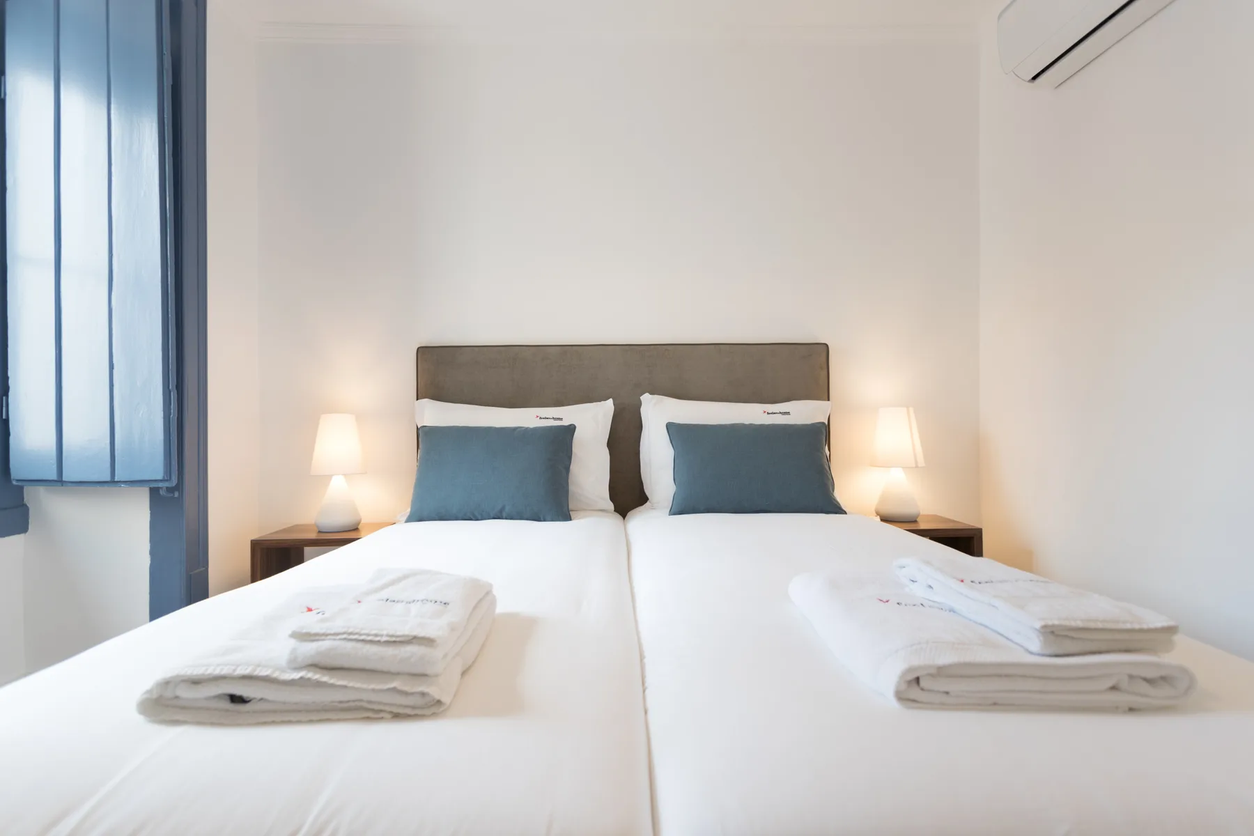 Un dormitorio luminoso con dos camas, lámparas de noche y toallas dobladas.