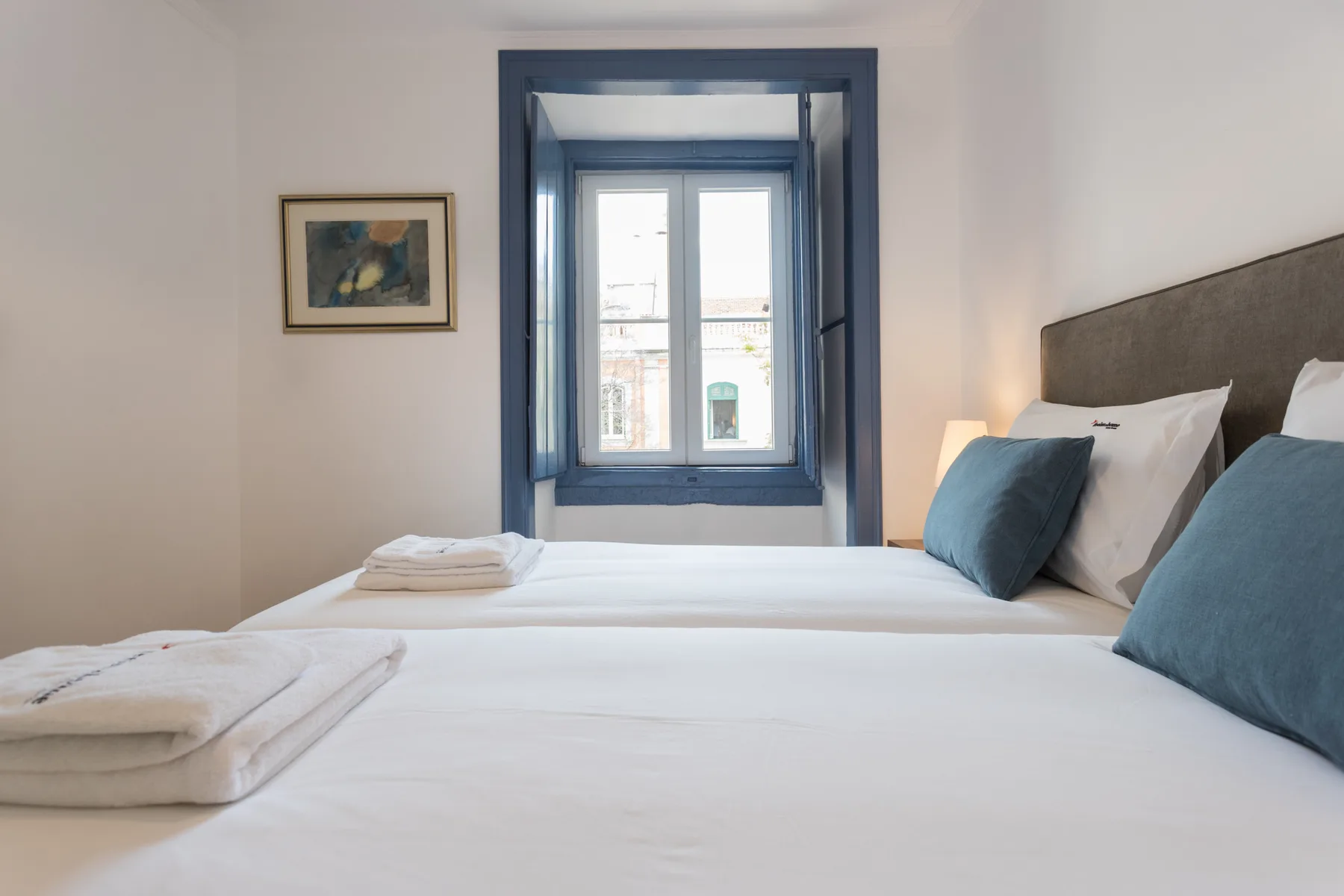 Dormitorio con ropa de cama blanca, almohadas azules y una ventana.