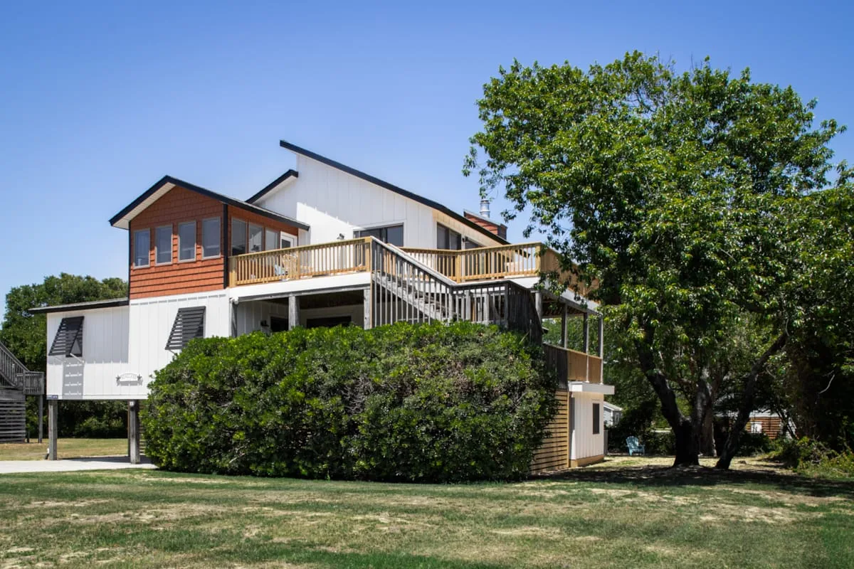 A raised house with a wooden deck stands beside a large leafy tree.