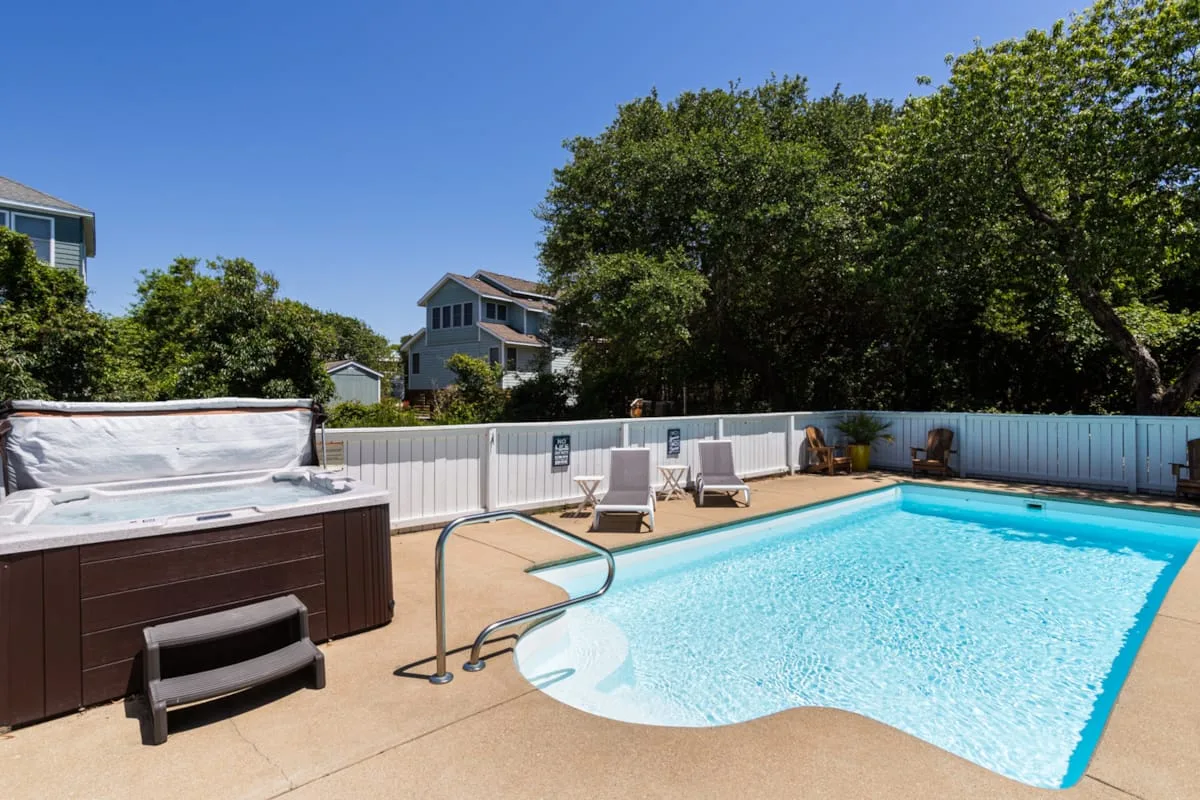 An outdoor swimming pool with lounge seating and trees behind a white fence.