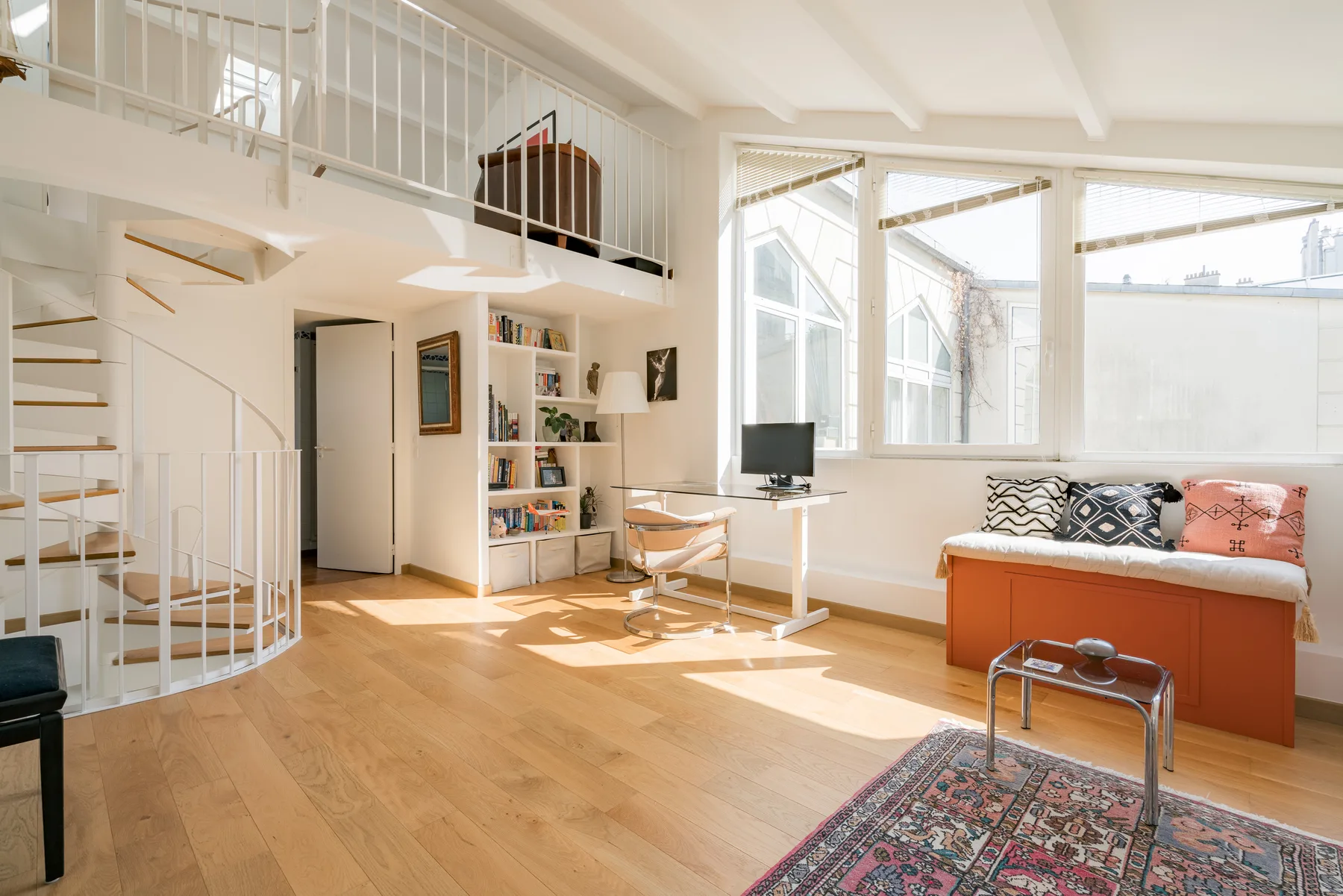 A bright loft living area with a staircase, desk, and seating by large windows.