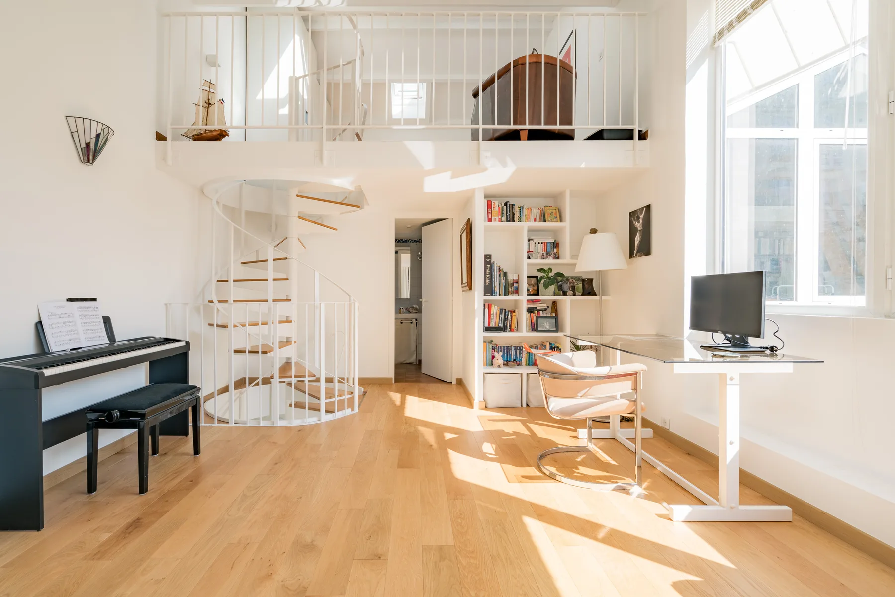A bright loft living area with a piano, desk, and spiral staircase.