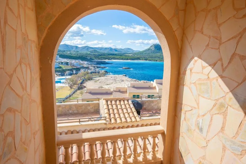 An arched balcony opening frames a coastal view of blue water and distant hills.