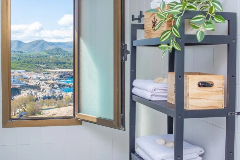 A bathroom with an open window overlooking a green valley and a storage shelf.