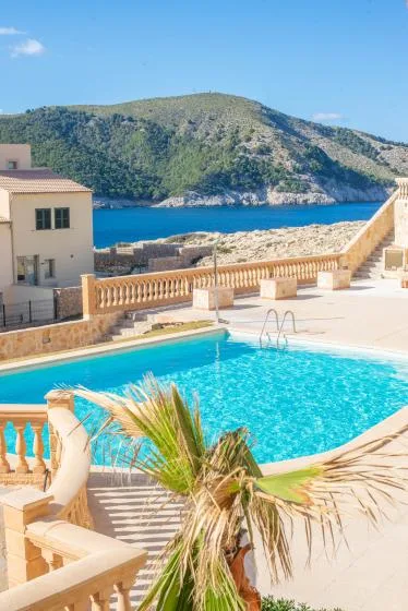 A swimming pool and terrace overlook blue water and a green hillside.