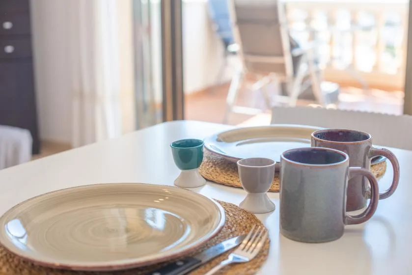 Dining table set with plates, cups, and cutlery beside a bright window.