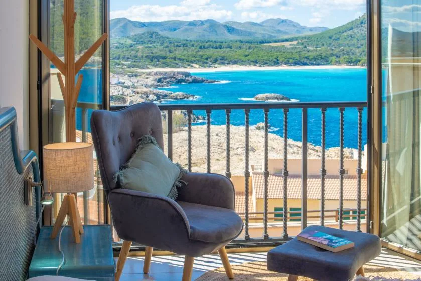A seating area beside an open balcony overlooks a bright blue bay and green hills.