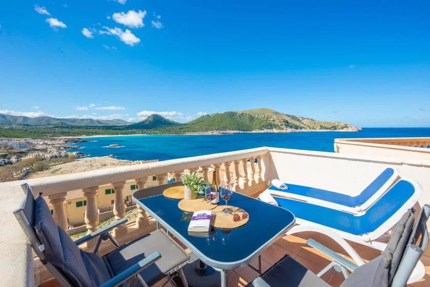 A seaside terrace overlooks the blue water, with outdoor seating and a dining table.