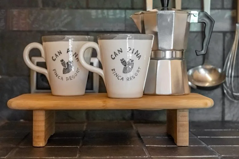Two mugs and a metal coffee maker sit on a wooden stand in a tiled kitchen.