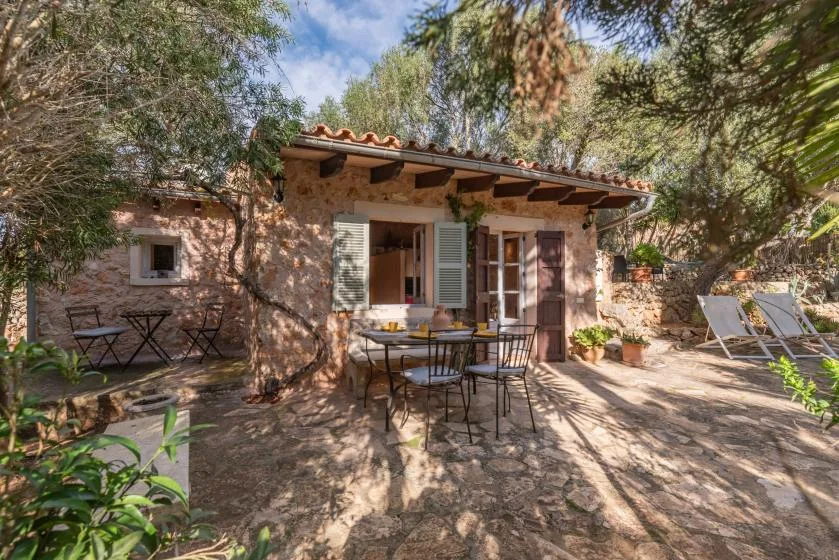 A stone house opens onto a shaded patio with outdoor seating and potted plants.
