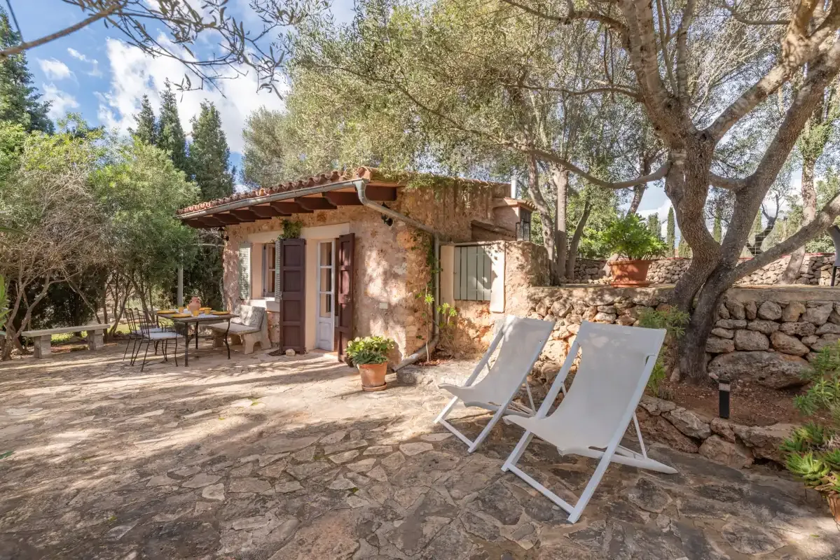 A small stone building opens onto a sunny patio with outdoor seating beneath trees.