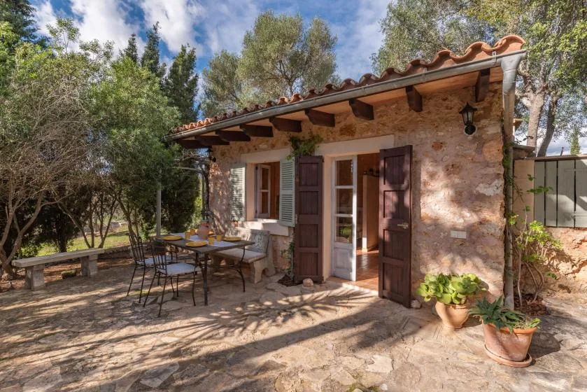 A stone cottage opens onto a sunny patio with outdoor seating and potted plants.