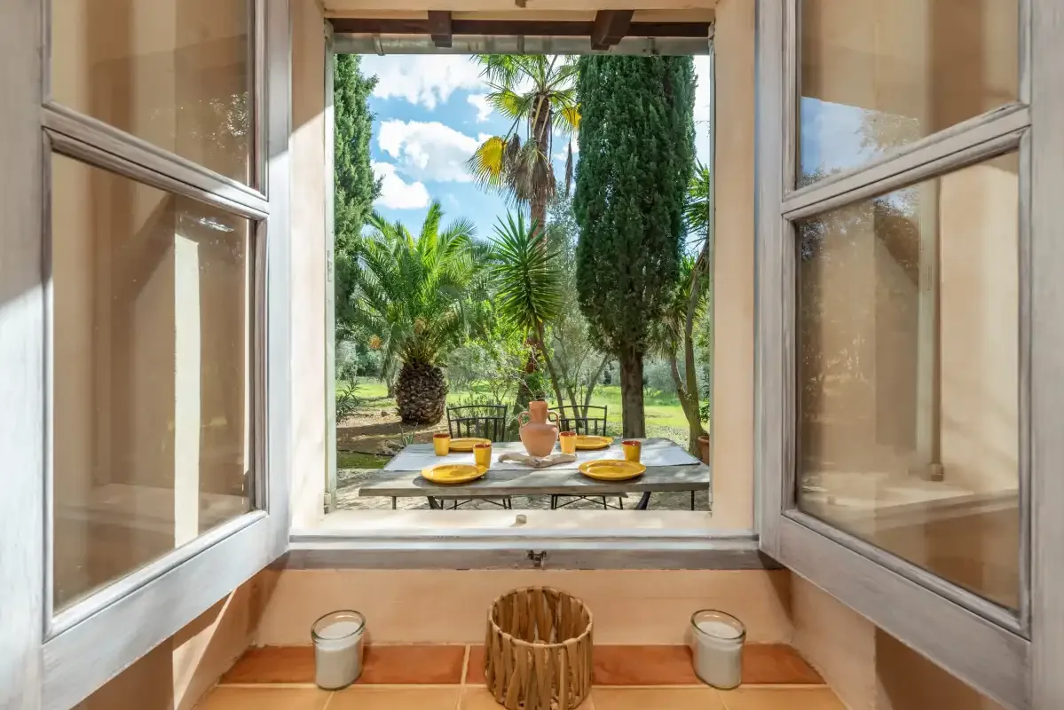 An open window overlooks a garden patio with table seating and palm trees.