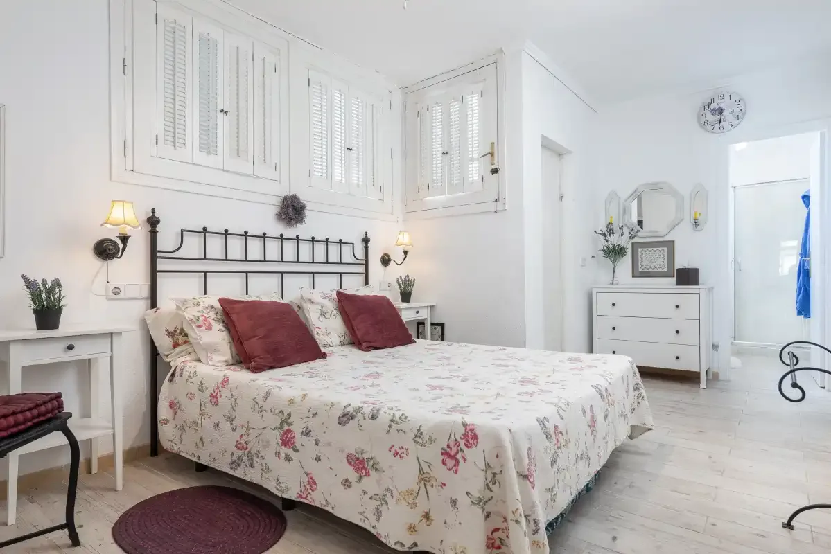 Un dormitorio luminoso con una colcha floral y contraventanas blancas.