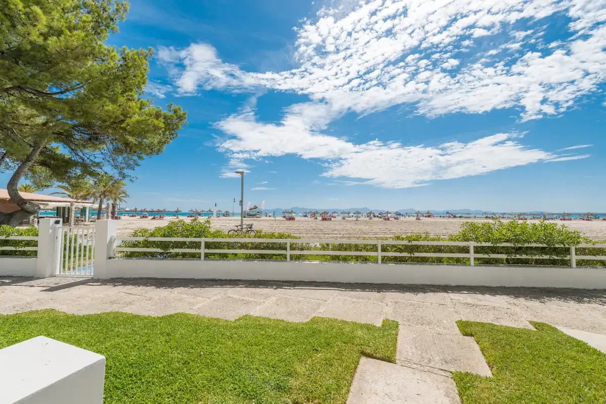 Vista junto al mar desde un jardín, con una valla blanca y la playa al fondo.