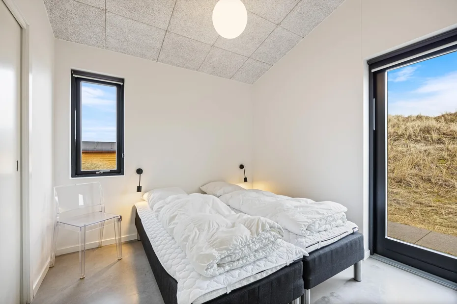 Dormitorio con una cama cubierta de ropa de cama blanca y ventanas con vistas a dunas cubiertas de hierba.
