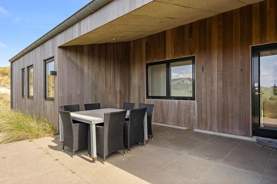 Comedor exterior cubierto con mesa y asientos junto a un edificio revestido de madera.