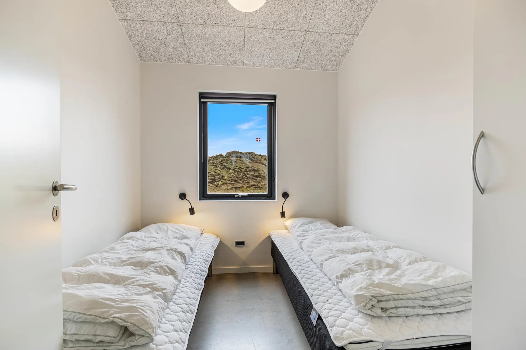 Un dormitorio luminoso con dos camas individuales frente a una ventana con dunas cubiertas de hierba.