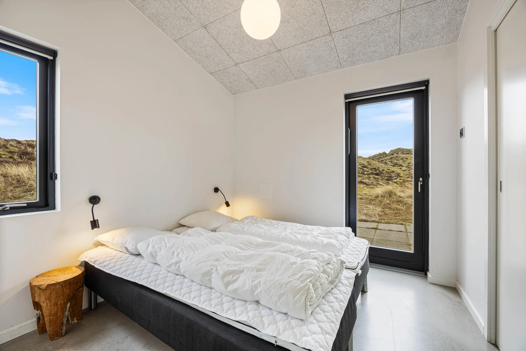 Un dormitorio luminoso con una cama junto a ventanas que dan a dunas cubiertas de hierba.