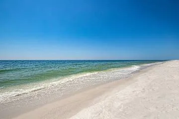 Una playa de arena se encuentra con aguas tranquilas azul verdosas bajo un cielo despejado.